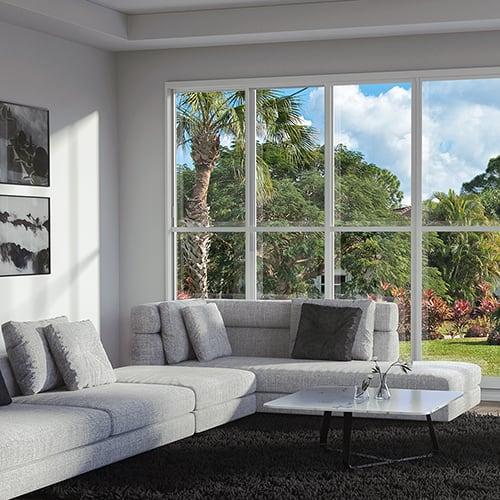 A point of view from inside a Florida room that also shows the outside window view a backyard with palm trees and flowers. Inside has two pictures hanging on white walls, a floor covered by a black shag rug with a white table placed on top, next to gray couches with matching pillows.
